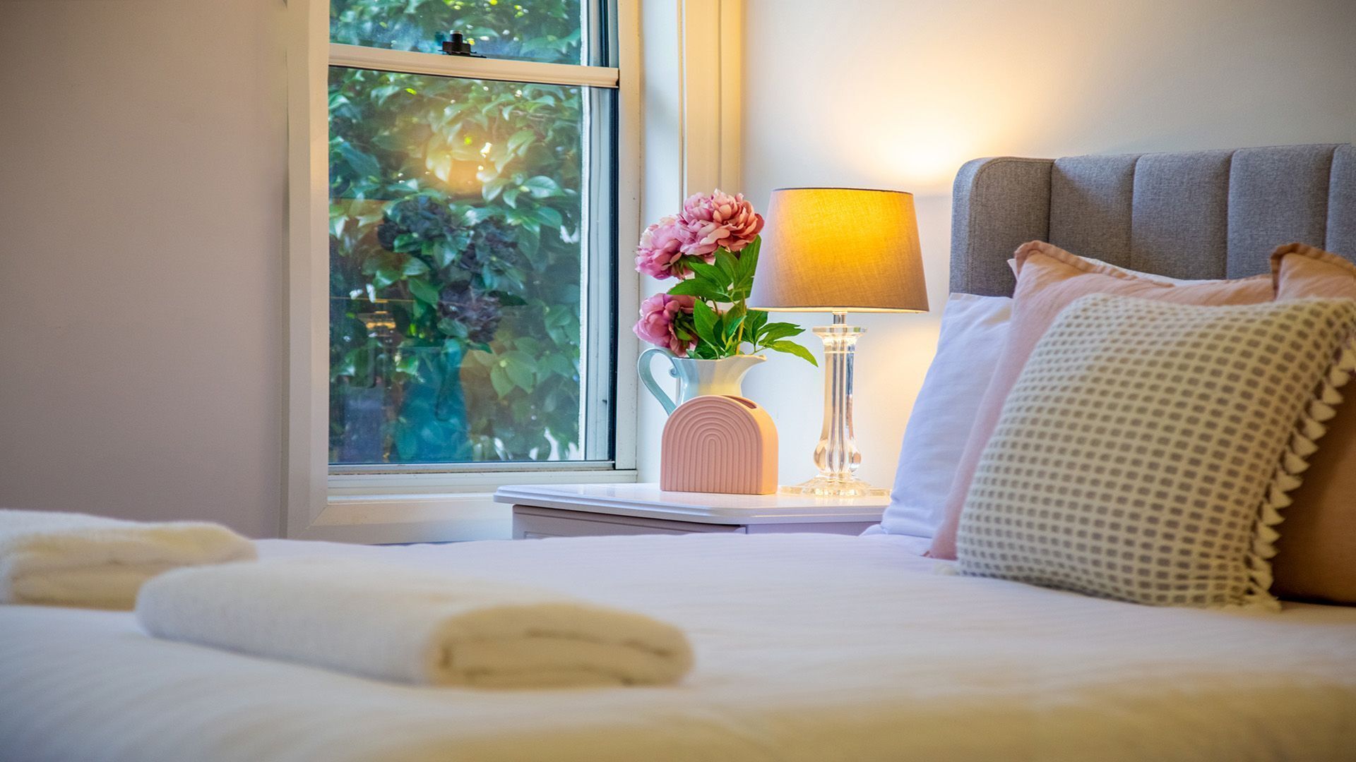 A neatly made bed with white linens, a bedside lamp, a vase with flowers, and a window with a view of greenery.