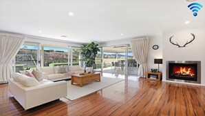 Amplia sala de estar con sofá blanco, mesa de centro de madera y una planta grande. Cuenta con chimenea, ventanas amplias y vistas al exterior.