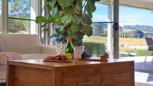 Una mesa de centro de madera con una botella, vasos y aperitivos en una habitación con un ventanal grande y una planta.
