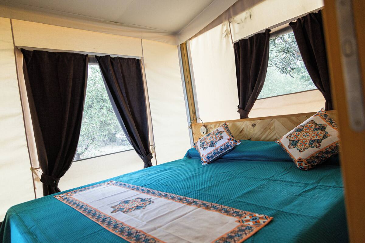 Un lit recouvert d’une couvre-lit turquoise et agrémenté de coussins décoratifs, une fenêtre avec des rideaux et une vue sur la verdure à l’extérieur.