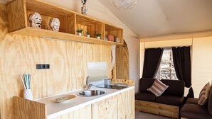 A compact kitchen with a wooden countertop, a sink, and a built-in oven. There is a wooden shelf above the counter with decorative items. A brown sofa with striped pillows is visible in the background. - Koru (Belpasso)