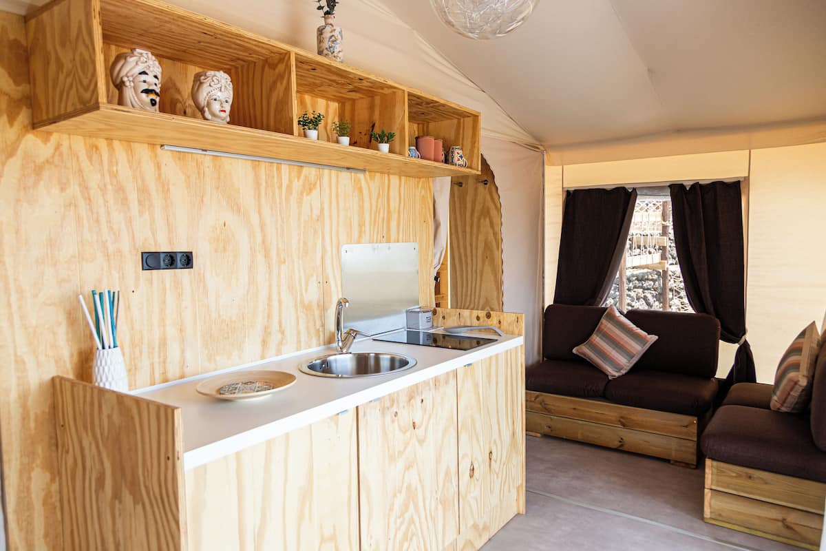 Une cuisine compacte avec un plan de travail en bois, un évier et un four encastré. Une étagère en bois, ornée d’objets décoratifs, se trouve au-dessus du plan de travail. On aperçoit un canapé marron avec des coussins à rayures à l’arrière-plan.