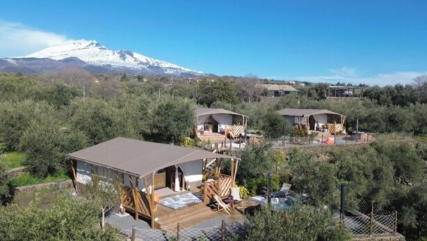 A mountain lodge with multiple cabins, each featuring a deck and outdoor seating area, surrounded by dense vegetation and a snow-capped mountain in the background. - Koru (Belpasso)