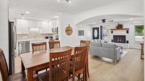 Une cuisine moderne avec des meubles blancs, un coin repas doté d’une table et de chaises en bois, un canapé gris et une cheminée.