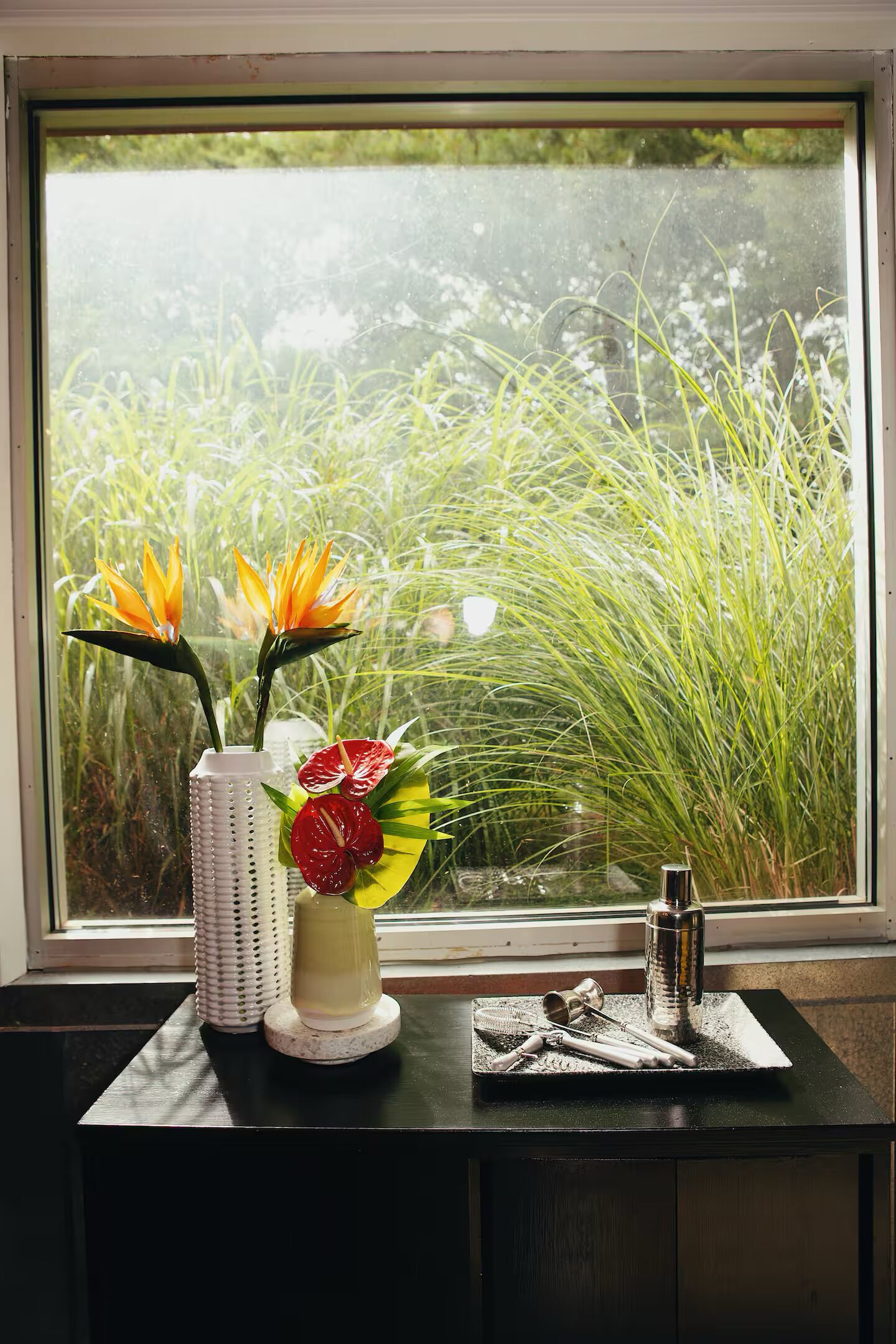 A window with a view of greenery, a white vase with flowers, and a small tray with grooming tools.