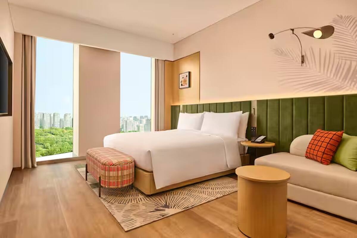 Ein modernes Hotelzimmer mit einem großen Bett, einem grünen Stoffpolster-Kopfende, einer kleinen Couch, einem runden Beistelltisch und Blick auf die Stadt.