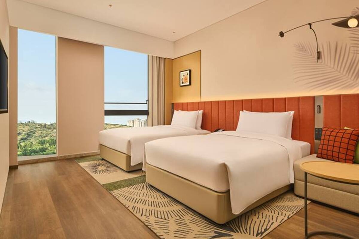 Ein Hotelzimmer mit zwei Betten, einem kleinen Tisch, einem Sessel, einem großen Fenster mit Aussicht und einer Deckenleuchte.