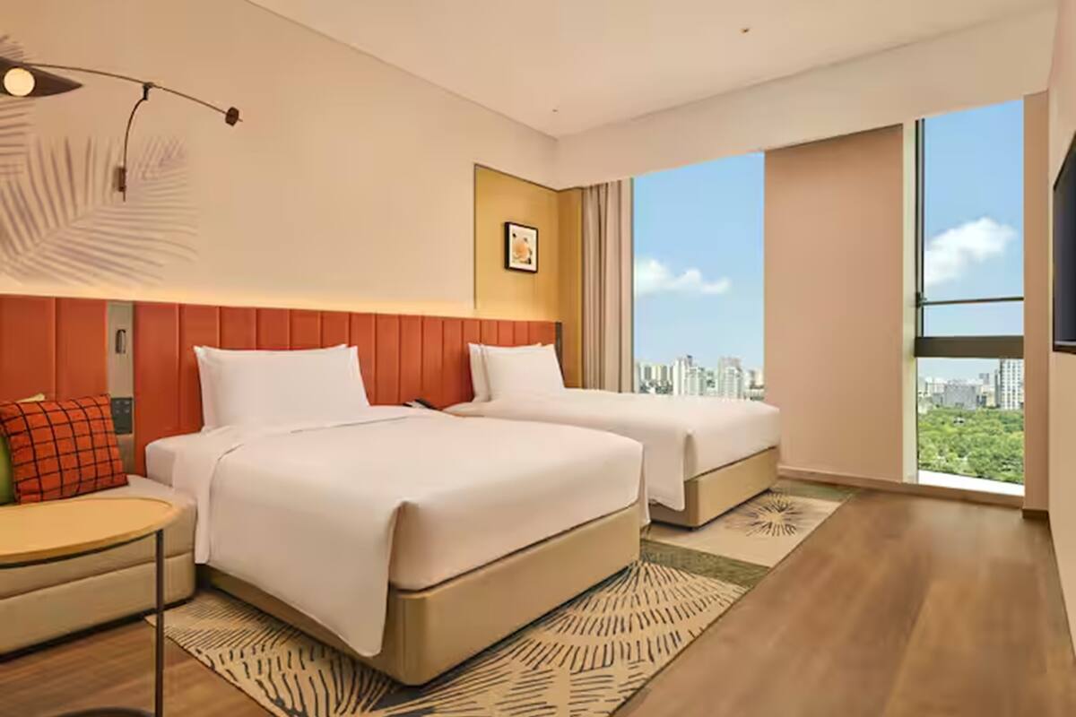 Ein Hotelzimmer mit zwei Betten, einem Nachttisch, einem großen Fenster mit Blick auf die Stadt und einer modernen Lampe.