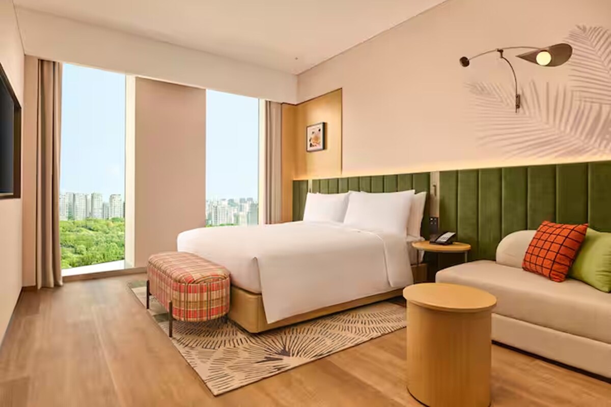 A modern hotel room with a large bed, a green upholstered headboard, a small sofa, a round side table, and a view of the city.