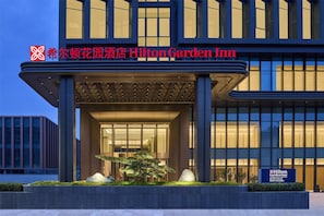 L’entrée d’un hôtel moderne se distingue par une grande façade vitrée, un hall d’entrée bien éclairé et un jardin aménagé avec un arbre remarquable.