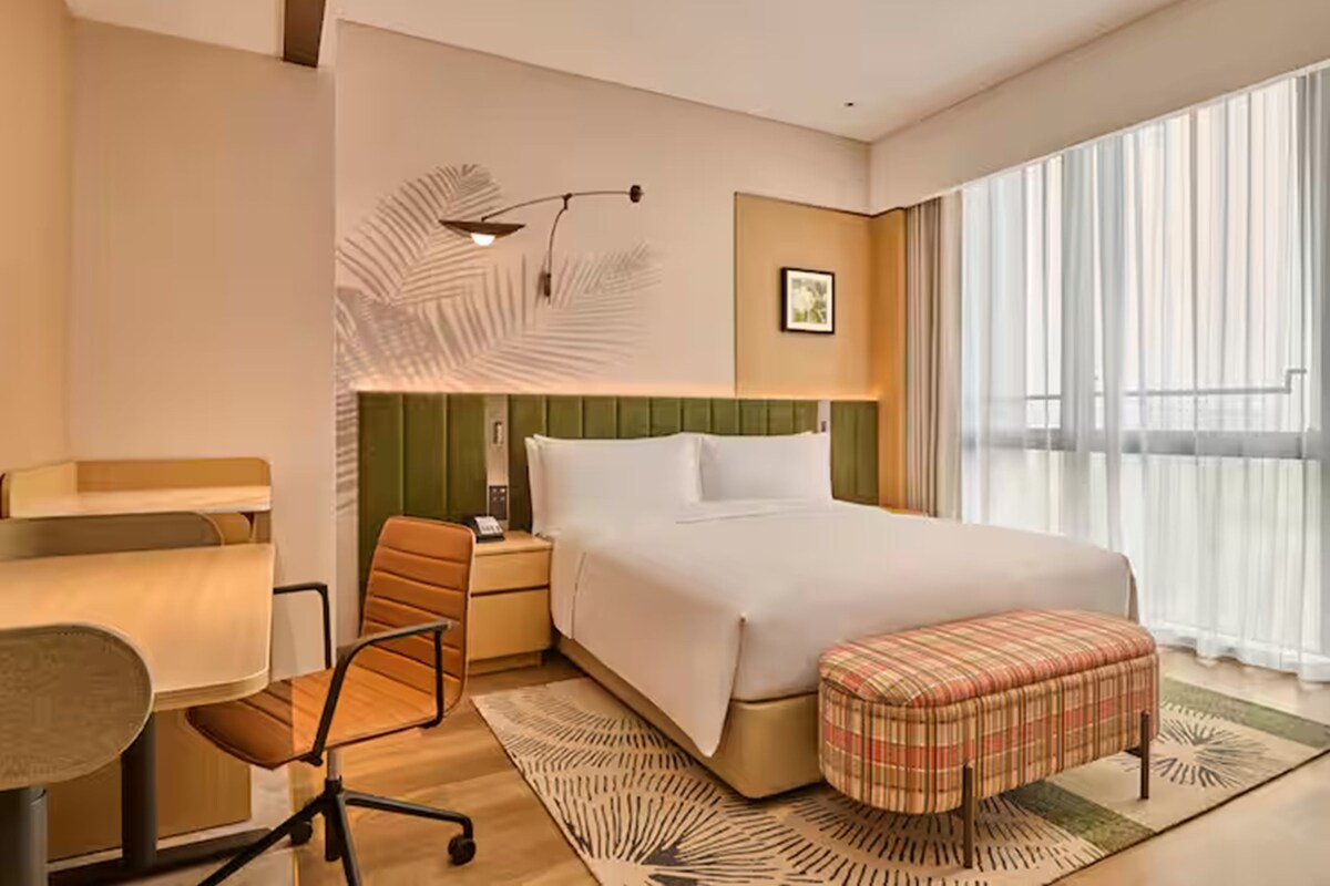 A hotel room with a bed, a desk, a chair, a nightstand, a lamp, and a large window with curtains.