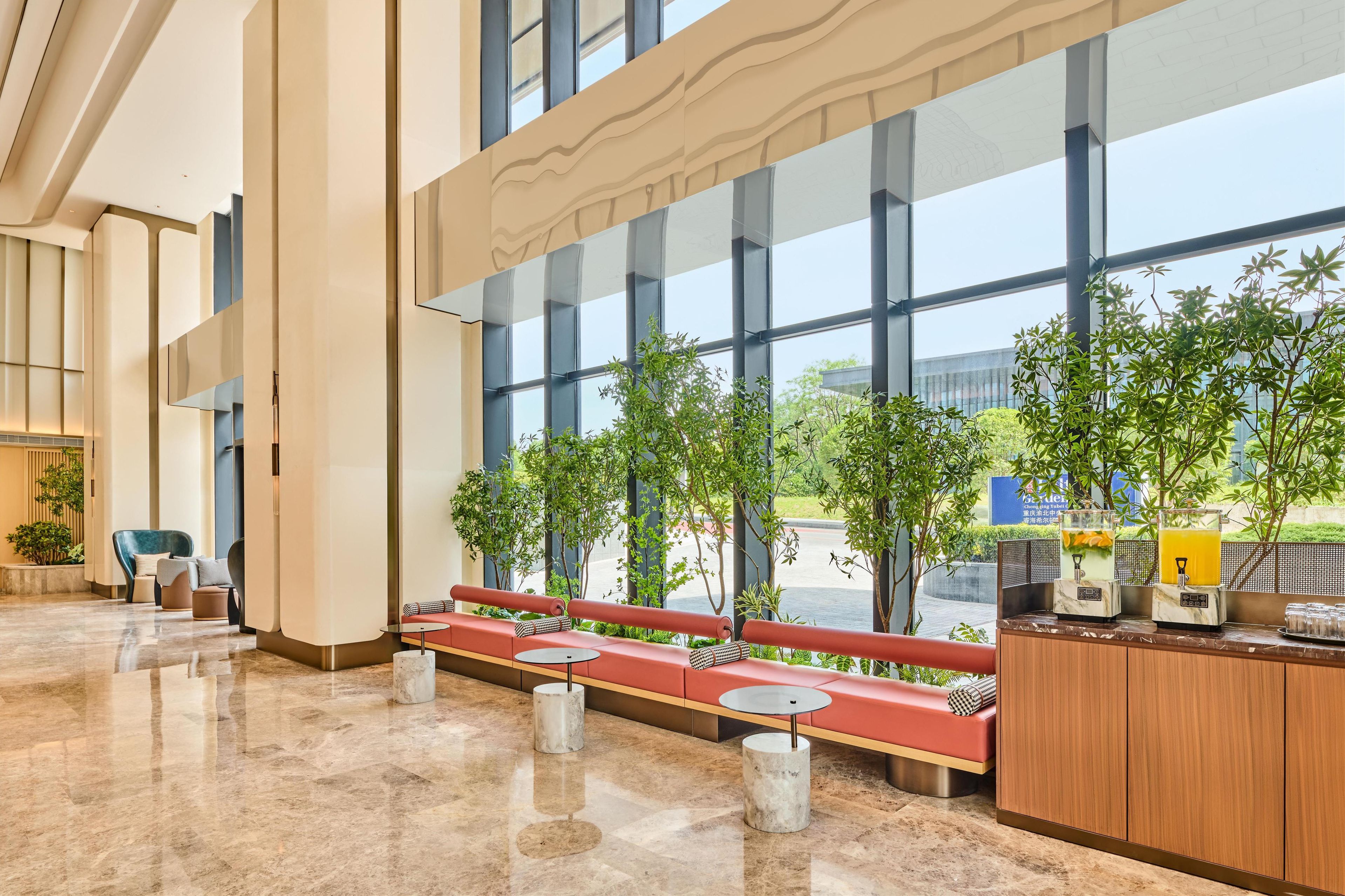A spacious lobby with a long red bench, marble flooring, and large windows offering views of greenery and a building in the background.