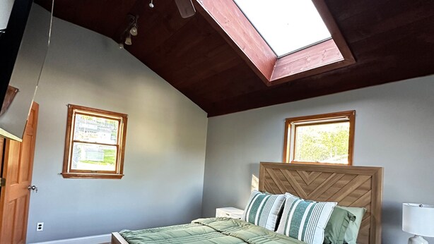 Un dormitorio con techo inclinado, una cama con cabecera de madera, una mesita de noche con lámpara y dos ventanas con persianas.