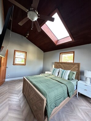 Un dormitorio con techo inclinado, una cama con cabecera de madera, una mesita de noche con lámpara y dos ventanas con persianas.