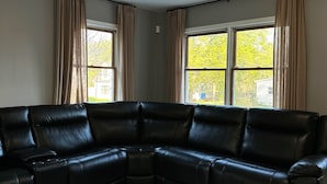 Una sala de estar con un sofá seccional de cuero negro, un ventilador de techo y un ventanal con cortinas.