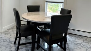 Un comedor con una mesa redonda, cuatro sillas, una lámpara de techo, una ventana con persianas y suelo de mármol.