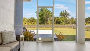 Una habitación con un amplio ventanal corredera que da a un jardín con césped, un banco acolchado y una silla de mimbre.