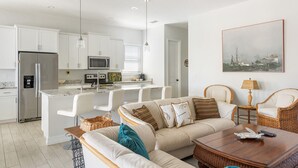 Una cocina moderna con armarios blancos, un refrigerador de acero inoxidable y un microondas integrado. Hay un comedor con sillas blancas y una mesa de madera. La sala de estar cuenta con un sofá color beige con cojines estampados y una mesa de centro de madera.