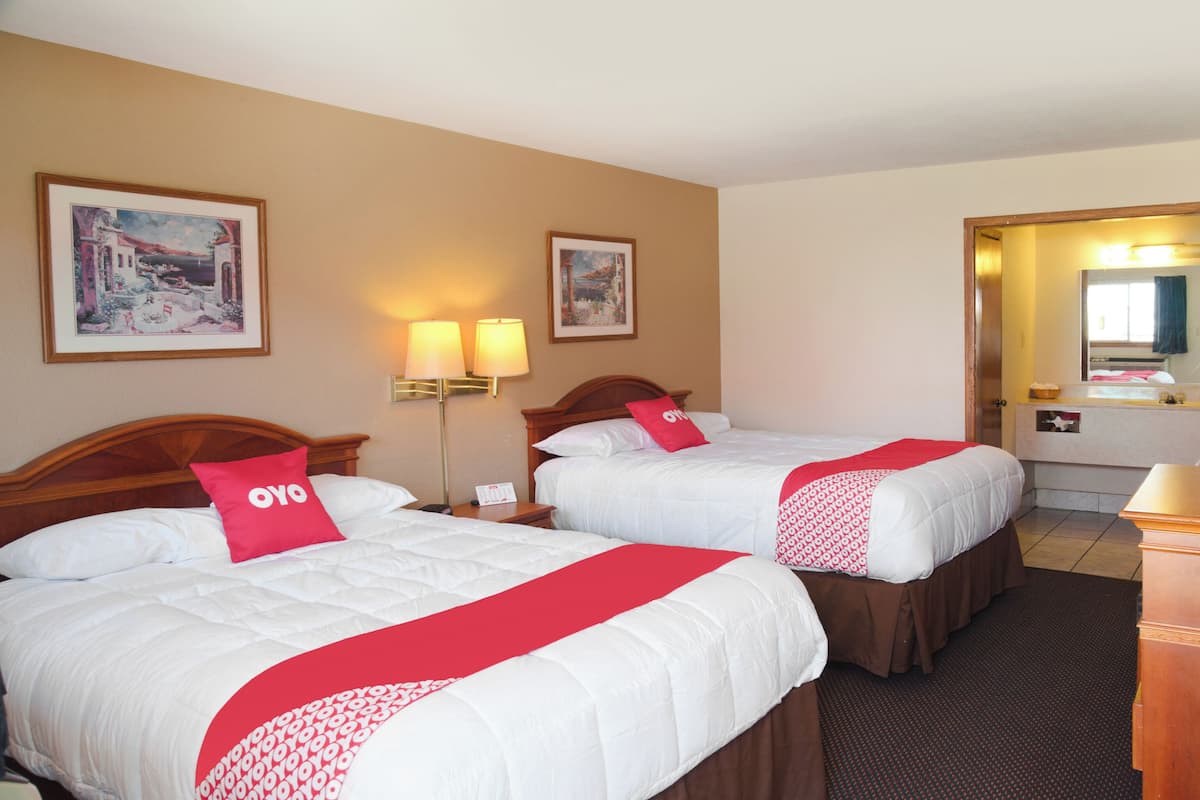 Une chambre d’hôtel avec deux lits, chacun agrémenté de draps blancs et de touches de couleur rouge, une table de chevet avec une lampe, et une salle de bain visible à travers une porte ouverte.