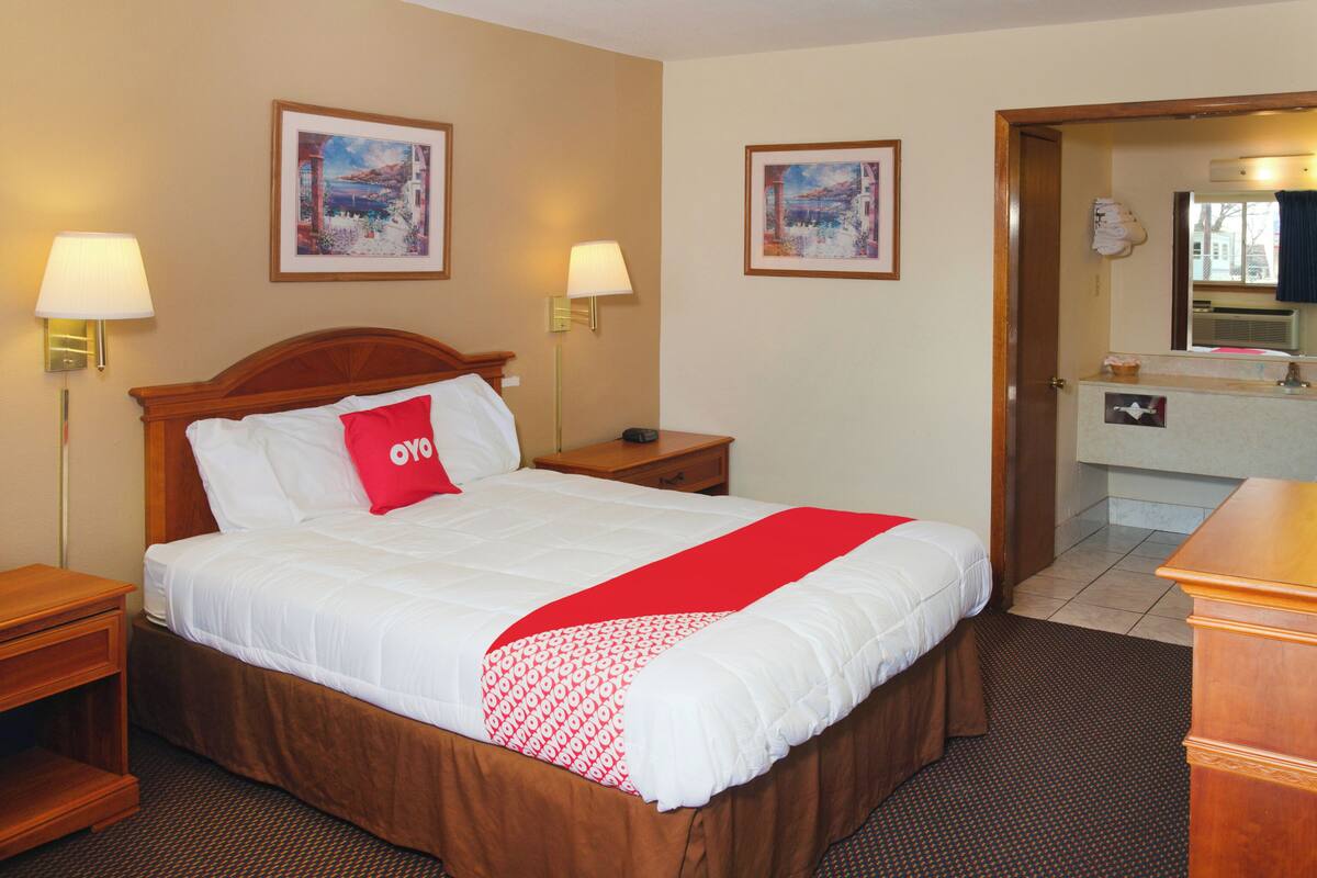 Une chambre d’hôtel avec un lit, deux lampes de chevet, une table de chevet, une tête de lit en bois, un coussin décoratif rouge, deux tableaux encadrés accrochés au mur, une salle de bain visible à travers une porte ouverte et un bureau en bois.