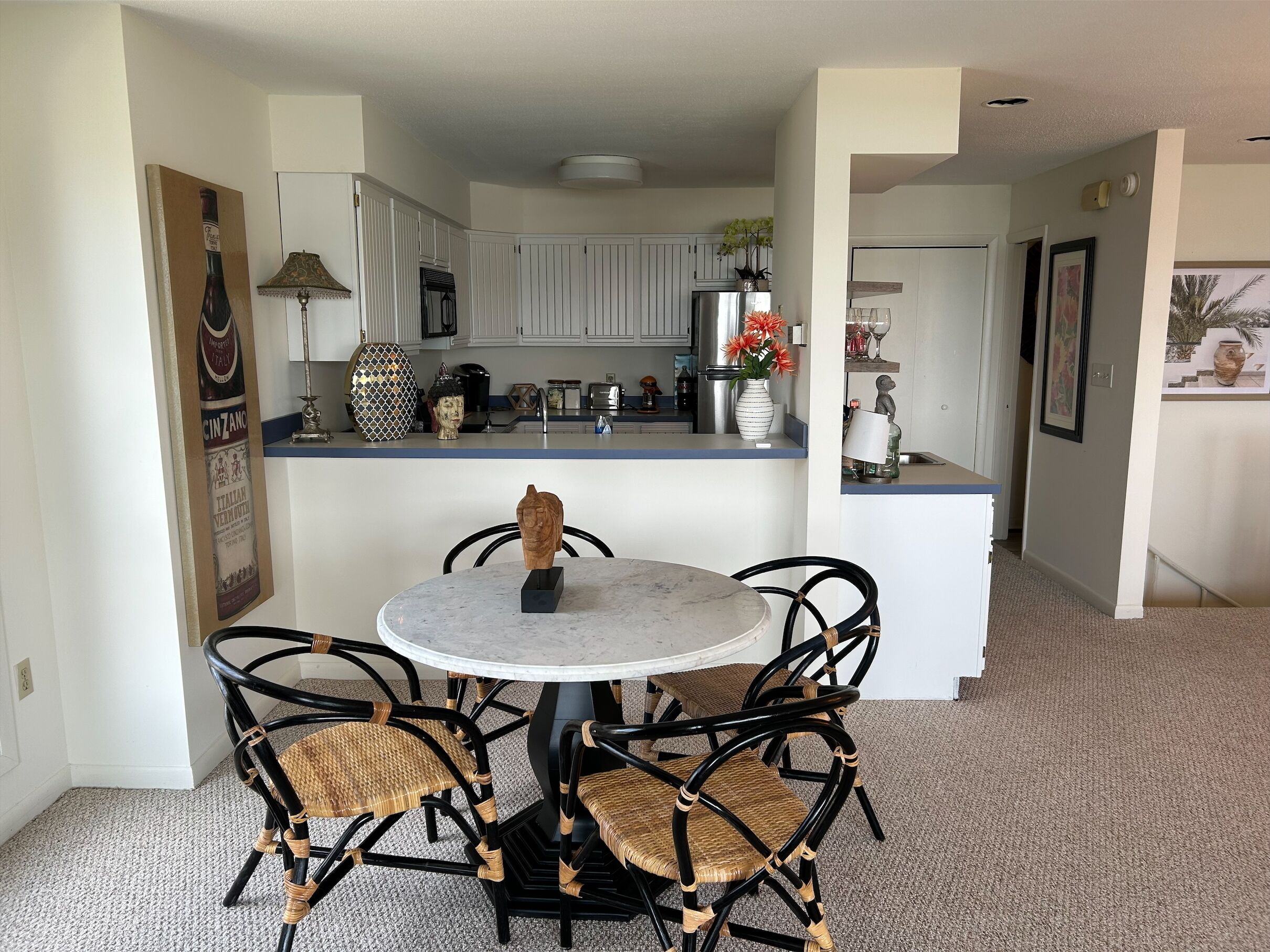 Un comedor con una mesa redonda y cuatro sillas de mimbre, una cocina al fondo y un pasillo que conduce a otra habitación.
