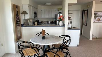 Un comedor con una mesa redonda y cuatro sillas de mimbre, una cocina al fondo y un pasillo que conduce a otra habitación.
