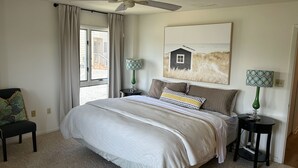 A bedroom with a bed, two bedside tables, a ceiling fan, a window with curtains, and a painting on the wall.
