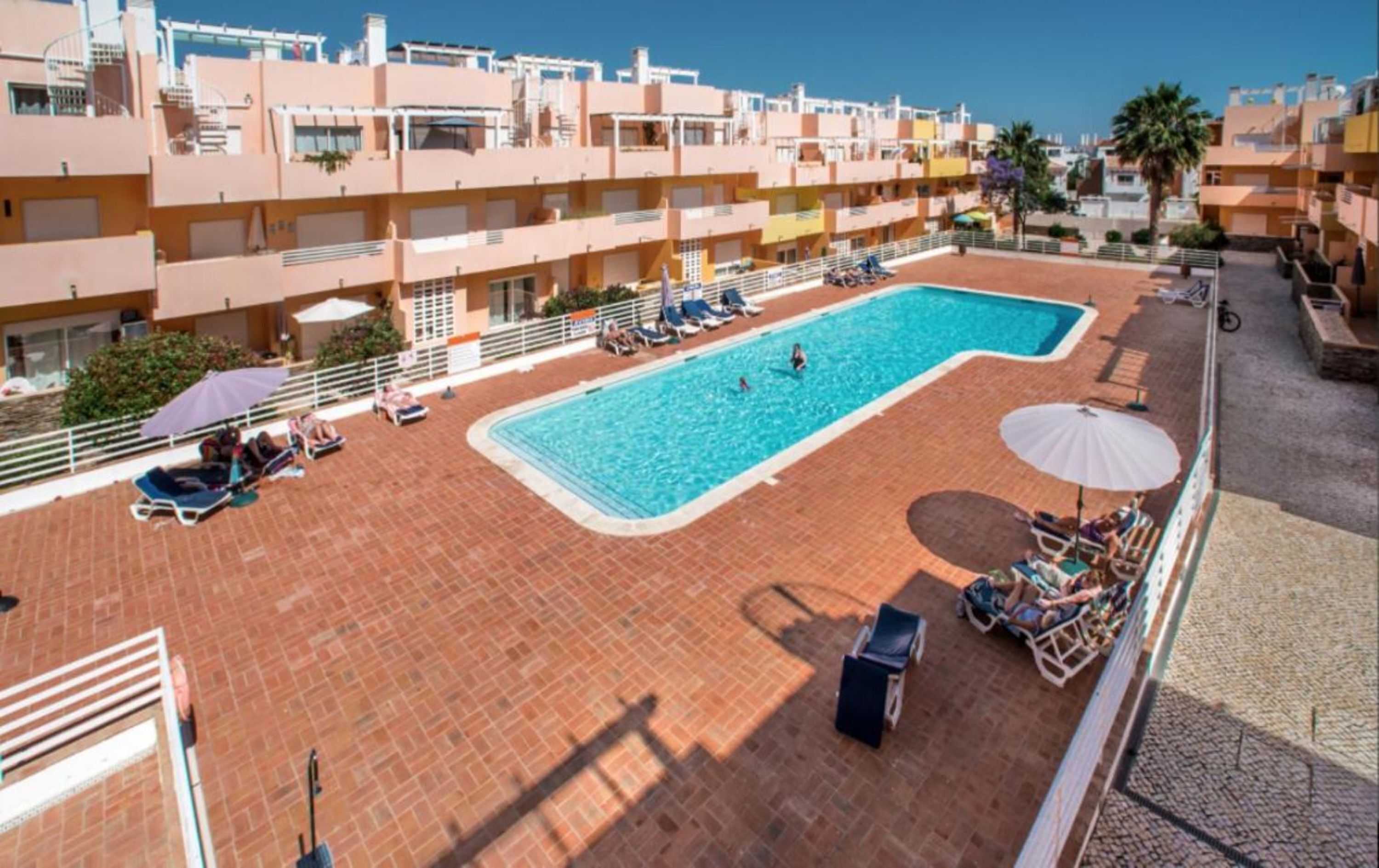 A rooftop swimming pool with sun loungers and umbrellas, surrounded by multi-story buildings.