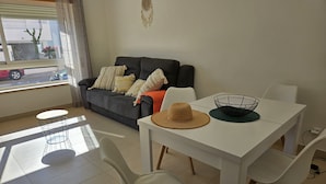 A living room with a grey sofa, white chairs, a white table, and a window with a view of the street. - Balcony Pool Royal Cabanas Golf 18 (Tavira)