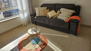 A living room with a grey sofa, patterned cushions, a dreamcatcher on the wall, and a window with a view of buildings. - Balcony Pool Royal Cabanas Golf 18 (Tavira)