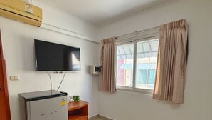 A room with a flat-screen TV mounted on the wall, a small refrigerator, a wooden cabinet, and a window with curtains. - Malee Guesthouse & Restaurant (Pattaya)