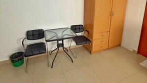 A room with a glass-top table, four black chairs, a wooden wardrobe, and a green trash bin. - Malee Guesthouse & Restaurant (Pattaya)