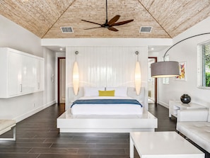A modern bedroom with a bed centered under a sloped ceiling, a ceiling fan, and a large window with blinds.