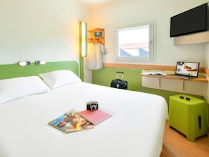 A hotel room with a bed, a desk with a laptop, a suitcase, and a magazine on the bed. - Ibis Budget Clermont Ferrand Sud Aubière (Aubiere)