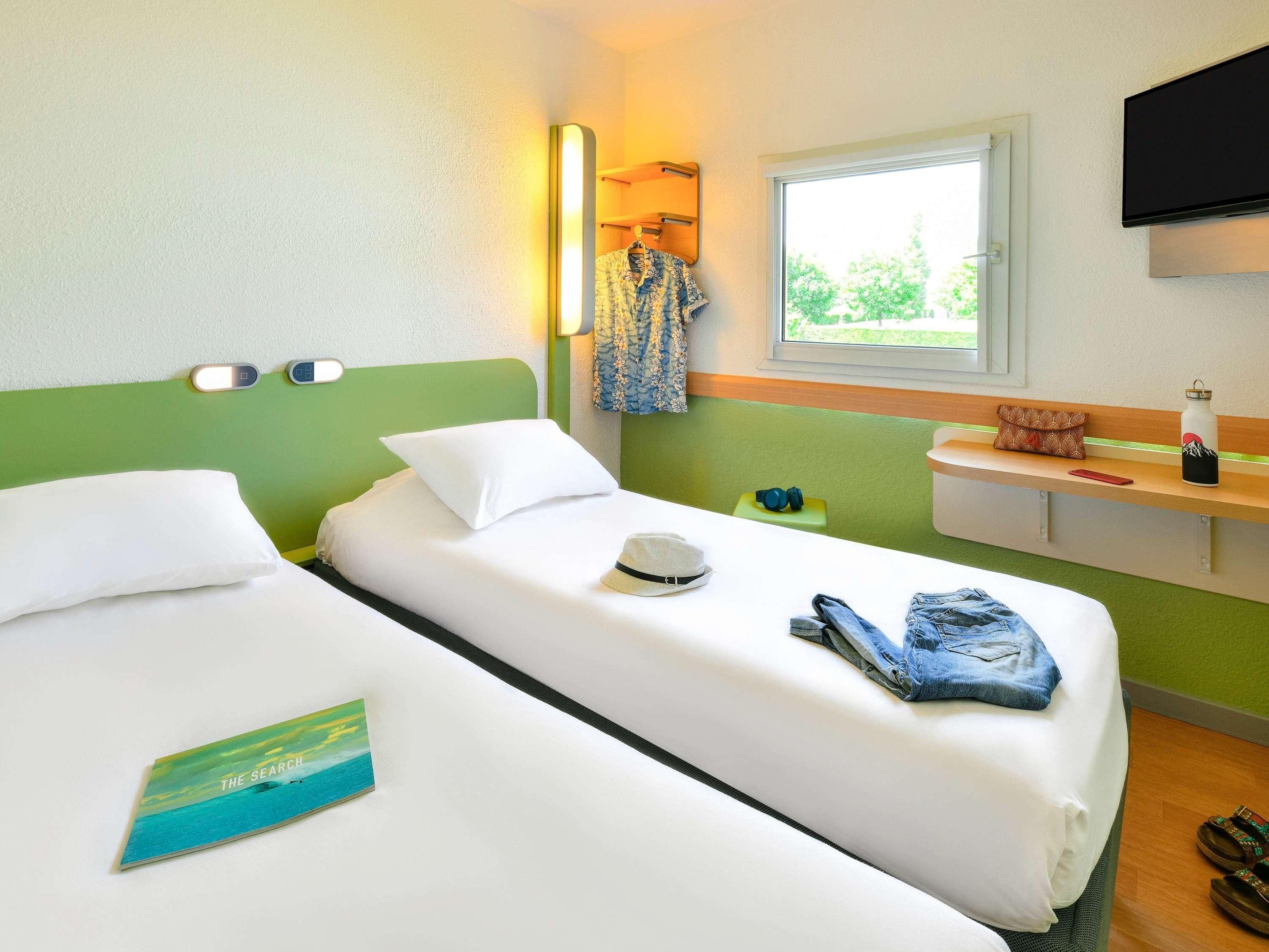 Une chambre d’hôtel avec deux lits, une fenêtre donnant sur un beau paysage, un téléviseur fixé au mur et une table de chevet avec une bouteille d’eau et un chapeau.