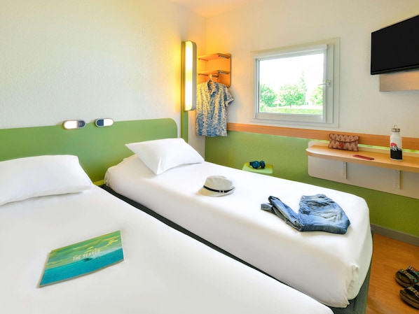 Ein Hotelzimmer mit zwei Betten, einem Fenster mit Blick, einem an der Wand montierten Fernseher und einem Nachttisch mit einer Wasserflasche und einem Hut.