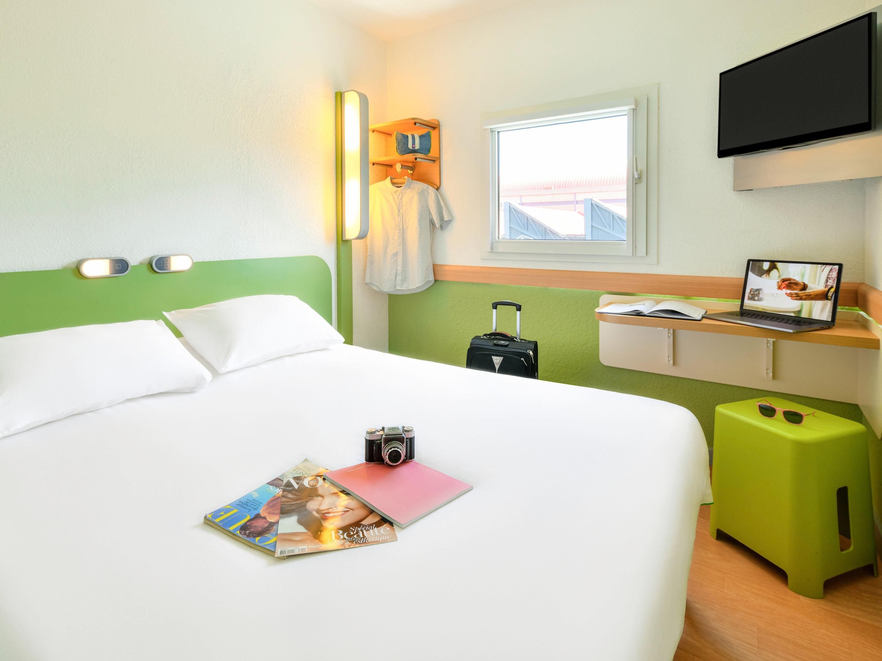 A hotel room with a bed, a desk with a laptop, a suitcase, and a magazine.