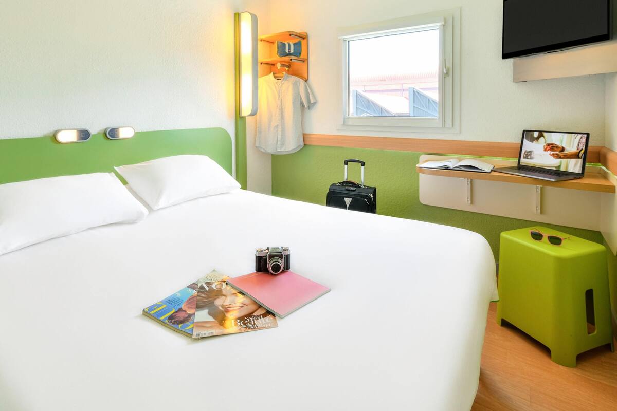 Una habitación de hotel con una cama, un escritorio, una computadora portátil, una maleta y una revista.