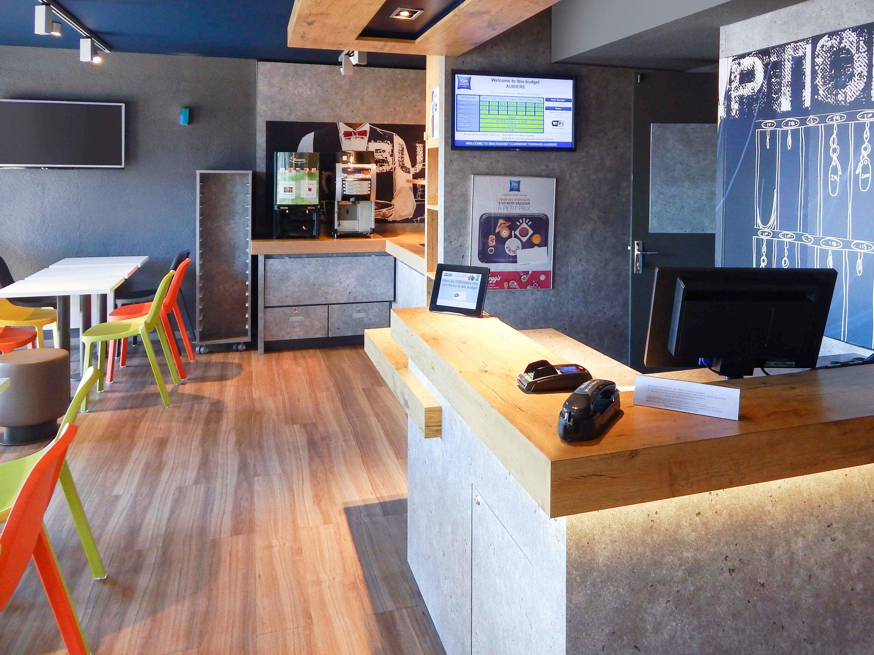 Un espace d’accueil moderne avec un comptoir en bois, un écran numérique affichant un message de bienvenue et des chaises colorées.