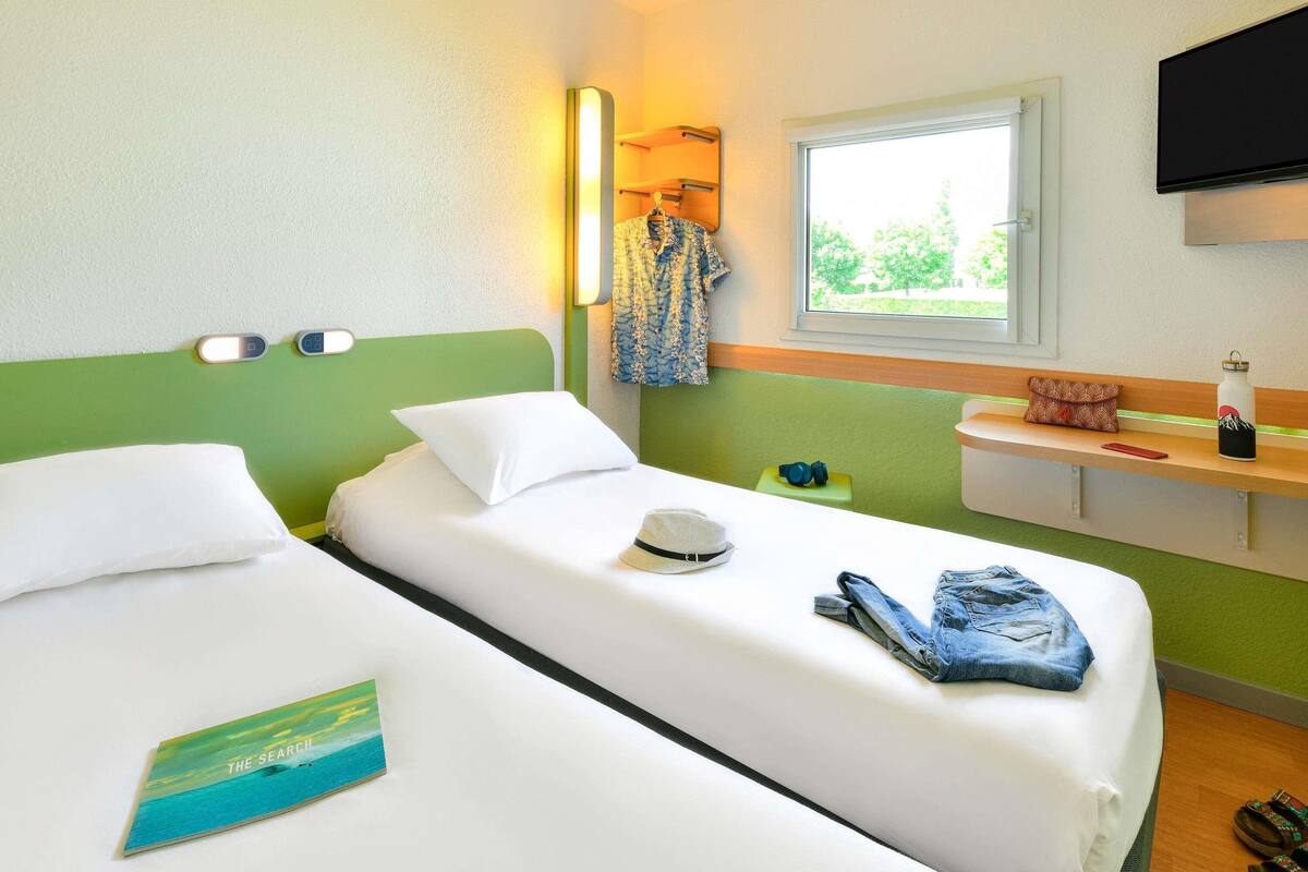 Habitación de hotel con dos camas individuales, una mesita de noche, un televisor y una ventana con vistas a zonas verdes.
