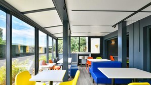 Een moderne, overdekte eetruimte met grote ramen, felgele en blauwe zitplaatsen en uitzicht op groen.