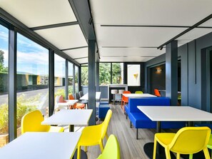 Un espace de restauration intérieur moderne, doté de grandes fenêtres, de sièges aux couleurs vives, jaune et bleu, et offrant une vue sur la verdure environnante.