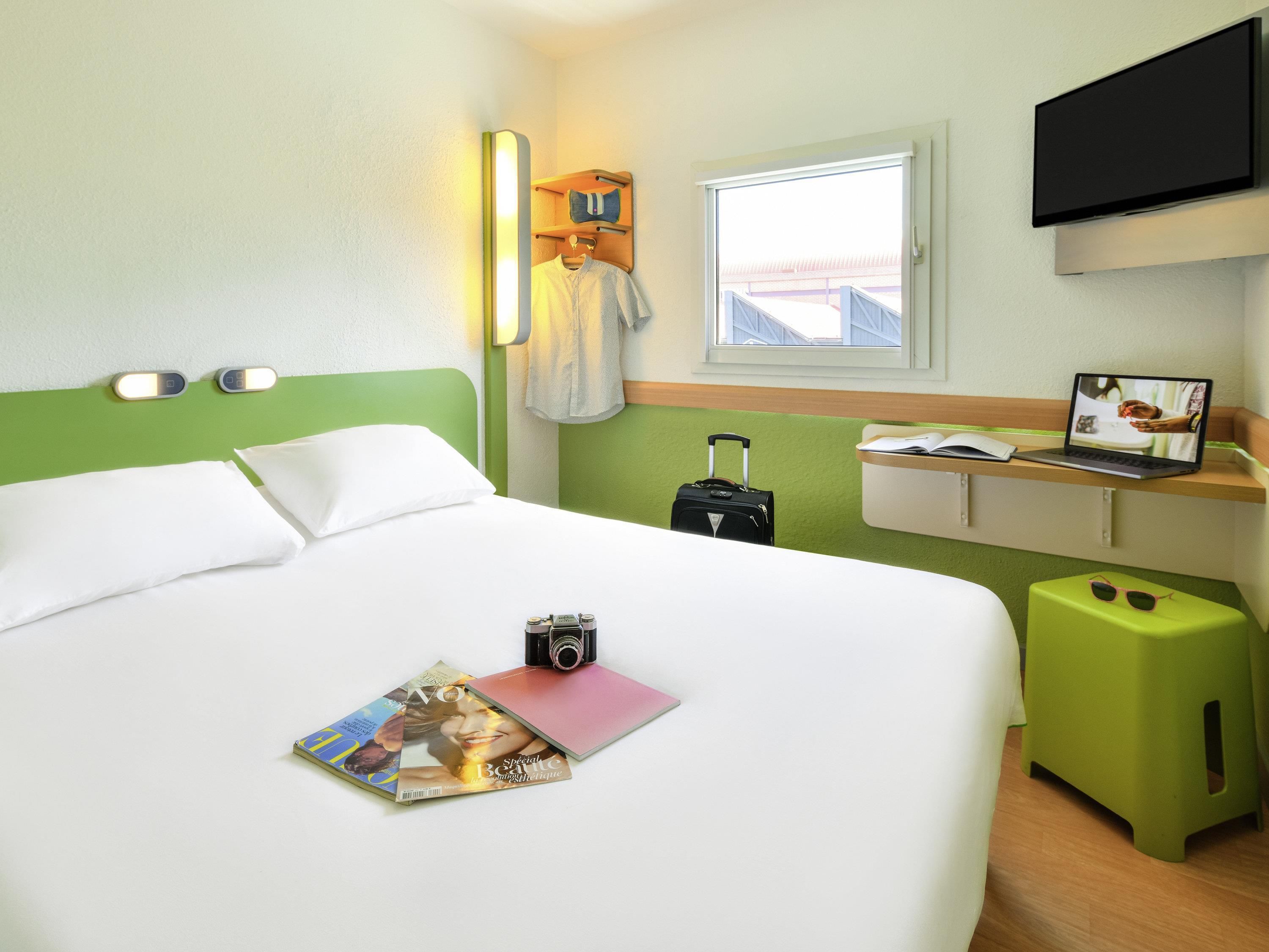 A hotel room with a bed, a desk with a laptop, a suitcase, and a magazine on the bed.