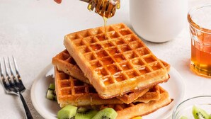 Een stapel wafels met honing die erover gedruppeld wordt, vergezeld van in plakjes gesneden kiwistukjes.