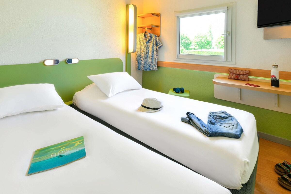 Une chambre d’hôtel avec deux lits simples, une table de chevet, une télévision et une fenêtre donnant sur un paysage verdoyant.