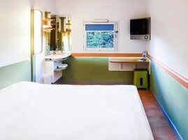 A small, single-room hotel room with a bed, a window, a sink, and a mirror.