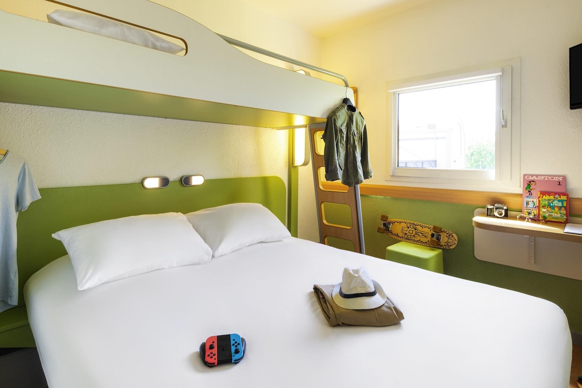 Una habitación de hotel con literas, una mesita con un monopatín y libros, y una ventana que deja entrar luz natural.