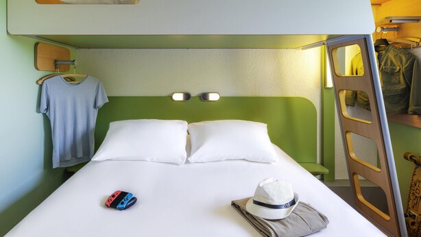 Een slaapkamer met stapelbedden, een groene muur, een houten ladder, een wit kussen, een hoed en een Nintendo Switch op het bed.