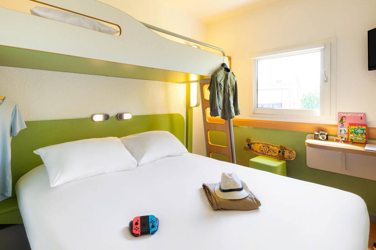 Una habitación de hotel con una cama litera, un pequeño escritorio con una cámara, un monopatín y un libro en el escritorio.