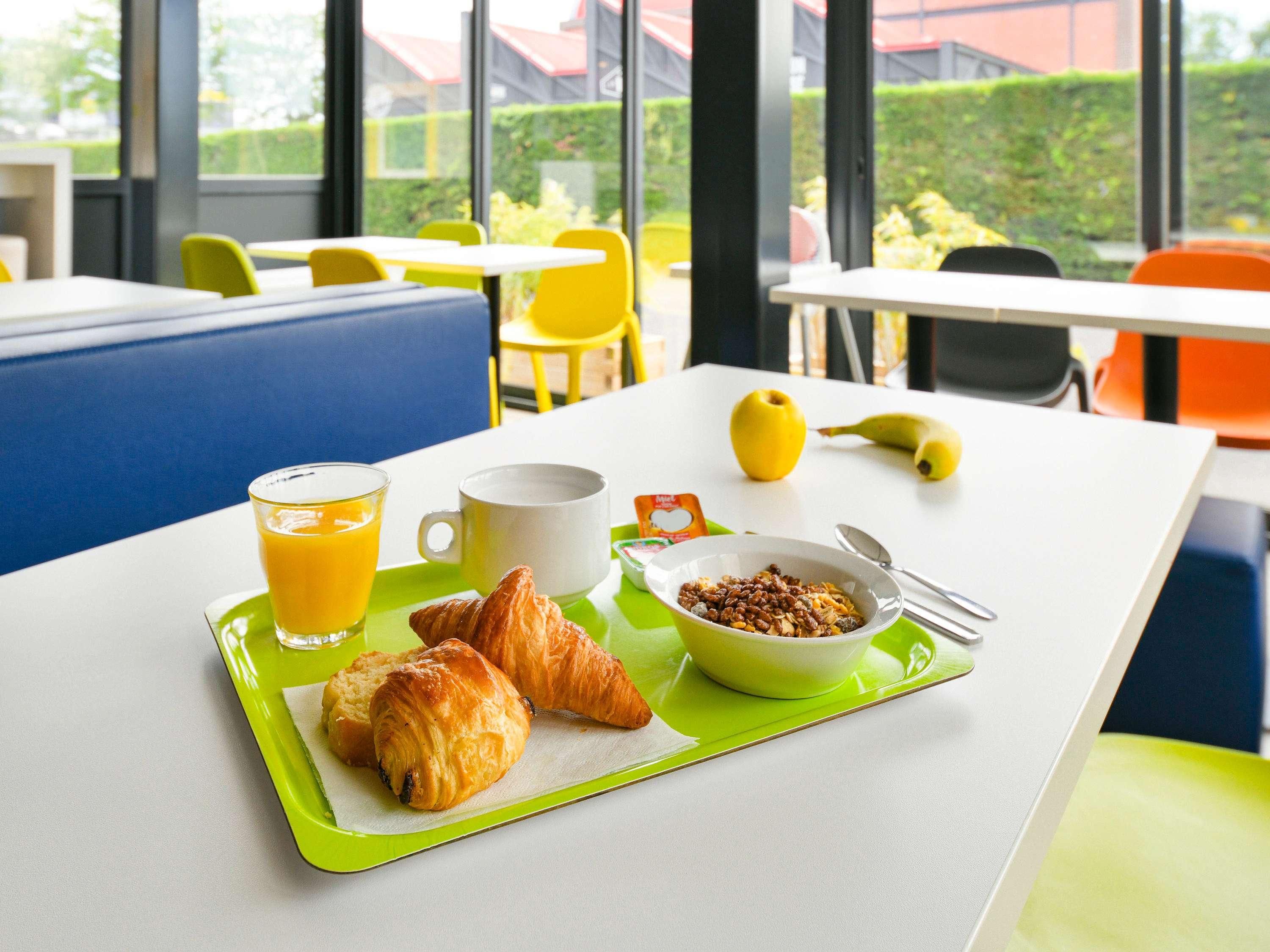 Bandeja de desayuno con cruasanes, un tazón de cereal, un vaso de jugo de naranja y un plátano sobre una mesa en una habitación luminosa con ventanas grandes y sillas amarillas.